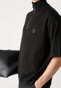 Černý pletený svetr s krátkým rukávem a polo zipem, který má texturovaný pruh. Na hrudi se nachází kulaté logo a hladká kožená taška.