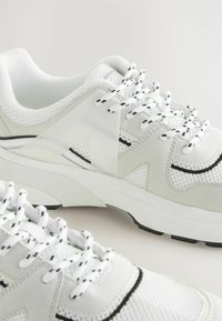 Baskets blanches de sport avec des matériaux en maille et synthétiques, ornées d'accents noirs, de superpositions texturées et de lacets contrastant noir et blanc.