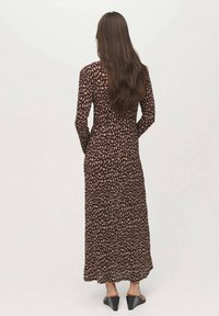 Robe longue marron à imprimé floral, avec un corsage ajusté et une jupe fluide. Manches longues et détail dans le dos visibles. Talons fins portés.