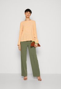 Haut à manches longues et ample en orange clair avec des motifs floraux blancs, associé à un pantalon large vert taille haute. Tenant une pochette marron.
