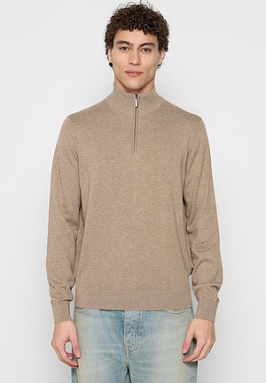 Junger Mann mit lockigem Haar trägt einen beigen Zip-up-Pullover und hellblaue Jeans und steht vor einem schlichten hellen Hintergrund.