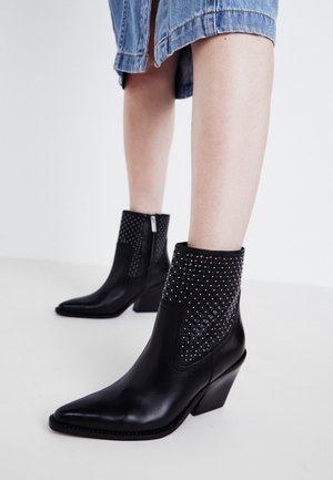 Botines negros de punta puntiaguda con detalles de tachuelas en la parte superior, usados con una falda de mezclilla sobre un fondo blanco liso.