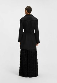 Black long coat with a broad collar, belted waist, and tailored fit. Paired with wide-leg black trousers featuring a textured, layered design.