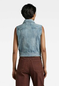 Ljusblå denim väst utan ärmar med krage, med sömnadsdetaljer och en kort design, som matchas med bruna byxor.