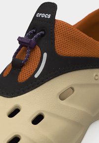 Κοντινό πλάνο ενός παπουτσιού Crocs σε καφετί και πορτοκαλί χρώμα με μαύρο περίγραμμα, μωβ ρυθμιζόμενα κορδόνια τύπου bungee και οπές εξαερισμού στη σόλα.