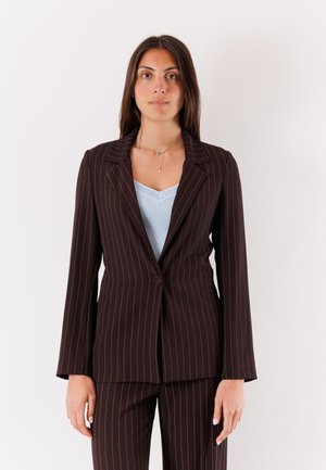 Donna con capelli lunghi e scuri che indossa un completo nero a righe sottili e una maglietta azzurro chiaro con bordi in pizzo, in piedi davanti a uno sfondo bianco uniforme.