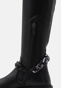 Botín negro de altura hasta la rodilla hecho de cuero sintético suave, con cremallera lateral, correa trenzada y detalle de cadena plateada brillante alrededor del tobillo.