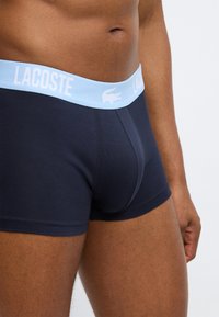 Shorts boxer bleu marine avec une ceinture élastique bleu clair portant le texte "LACOSTE" et le logo. Le tissu semble doux avec une coupe ajustée.