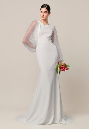 Naine elegantse valge kleidiga, läbipaistvate pärlitega kaunistatud patsmikega varrukatega, hoiab käes punaste tulpidega kimbu lihtsa beežika tausta ees.