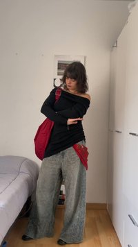 Person mit schulterlangem Haar, die ein schulterfreies schwarzes Oberteil, weite graue Hosen, schwarze Schuhe, eine rote Tasche und ein rotes Bandana trägt, in einem Schlafzimmer.