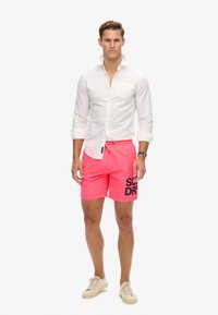 Mannenoutfit bestaande uit een wit overhemd met knopen en felle roze zwembroek met 'SUPERDRY' logo. Schoenen zijn lichtgekleurde sneakers.