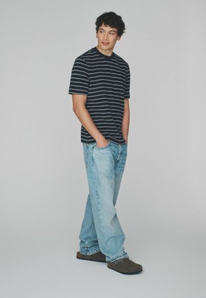 Junger Mann, der ein schwarz-weiß gestreiftes T-Shirt, hellblaue Jeans und dunkelbraune Slipper trägt, steht mit den Händen in den Taschen vor einem einfarbigen Hintergrund.