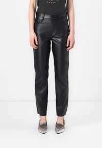 Femme portant un pantalon en cuir noir slim et des mocassins pointus argent métallisé, debout devant un fond blanc uni.