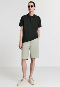 Uomo in maglietta polo nera, pantaloni beige, scarpe beige e occhiali da sole scuri, in piedi contro uno sfondo chiaro e uniforme.