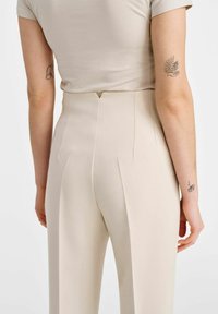 Beżowe dopasowane spodnie z zakładkami z tyłu, wysoki stan oraz gładka tekstura, w połączeniu z dopasowaną jasnobeżową bluzką.