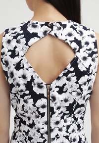 Bakvy av en ärmlös svart och vit blommig klänning med en diamantskuren utskärning, två knappar högst upp och en synlig dragkedja längs mitten.