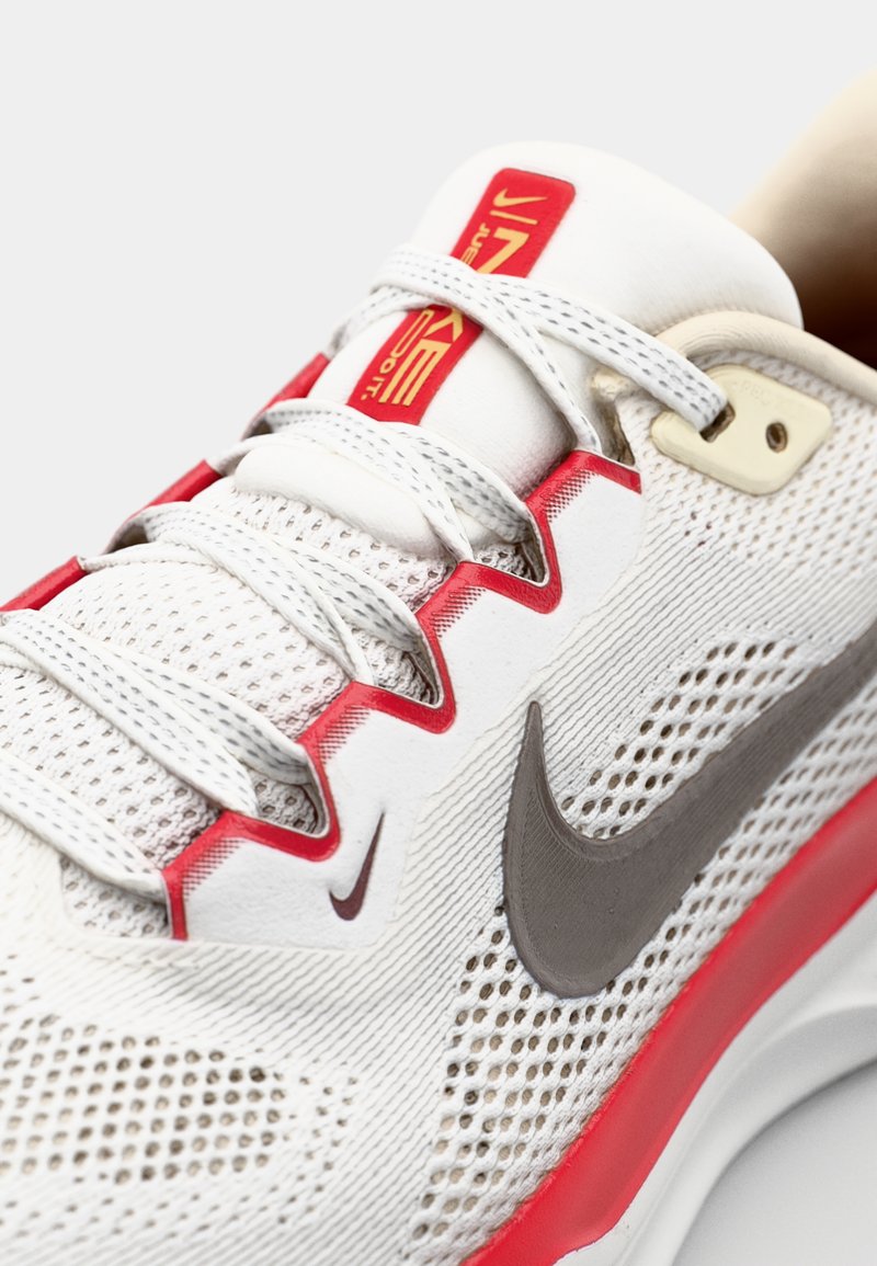 Scarpa sportiva bianca con parte superiore in mesh, dettagli rossi, logo Nike grigio, lacci testurizzati e colletto imbottito. Presenta il logo "Just Do It".