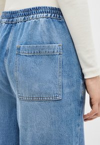 Nahaufnahme einer Person, die hellblaue Jeans aus Denim mit elastischem Bund und einer aufgesetzten Gesäßtasche trägt, wobei ein Teil eines cremefarbenen, gerippten Ärmels sichtbar ist.