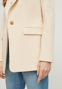 Femme portant un blazer beige à un bouton et poches à rabat, associé à un jean en denim bleu, la main reposant le long du corps.