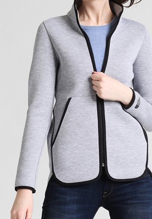 Persoon draagt een lichtgrijze jas met rits en zwarte bies over een blauw shirt, met één hand de jasrits vasthoudend.