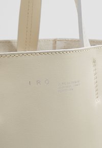 Beige läder tote bag med synlig sömnad, präglad logotyp och två remmar. Minimalistisk design med slät textur och inga ytterligare mönster.