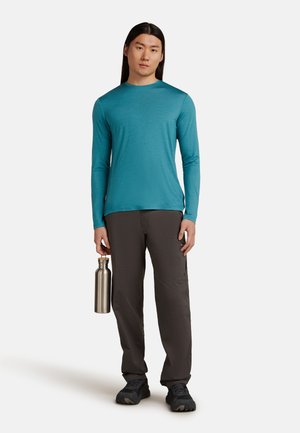 Langarmige teal shirt met ronde hals, gecombineerd met grijze broek en zwarte wandelschoenen. Houdt een zilveren metalen waterfles vast.