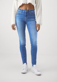 Högmidjade blå smala jeans med en blekt textur, femficksdesign och en guldknappsomkopplare, kombinerat med vita sneakers.