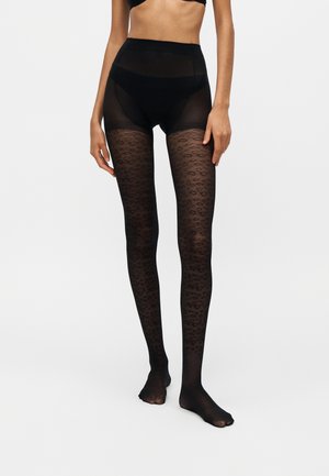 Femme portant des collants noirs transparents avec un motif léopard et un slip taille haute noir sur un fond uni.