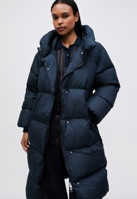 Žena ve dlouhém námořnickém péřovém kabátu přes tmavou košili na knoflíky a černé kalhoty, stojící na jednoduchém světlém pozadí.