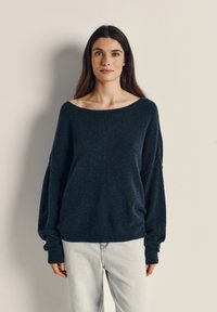 Marineblå oversized sweater lavet af blødt, struktureret stof med en bred halsudskæring og lange, tilspidsede ærmer, parret med lyse jeans.