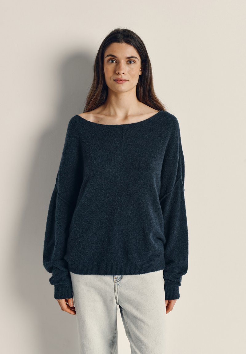 Marineblå oversized sweater lavet af blødt, struktureret stof med en bred halsudskæring og lange, tilspidsede ærmer, parret med lyse jeans.