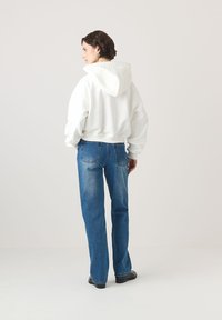 Biała krótkotnie z kapturem, w zestawieniu z niebieskimi dżinsami o prostym kroju. Tkanina wydaje się być miękka, a dżinsy mają lekki przetarcia i kieszenie z tyłu.