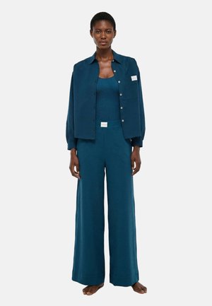 Femme debout pieds nus portant un pantalon ample vert sarcelle, un débardeur côtelé assorti et une chemise ouverte sur un fond uni.