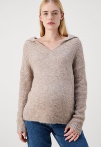 Pull en maille beige avec un col en V et un large col, manches longues et coupe décontractée. Texture douce et détail de l'ourlet côtelé. Porté avec un jean bleu.