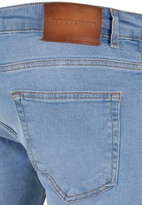 Jean en denim bleu clair avec une étiquette en cuir marron à la ceinture et des coutures orange le long de la couture et des poches.