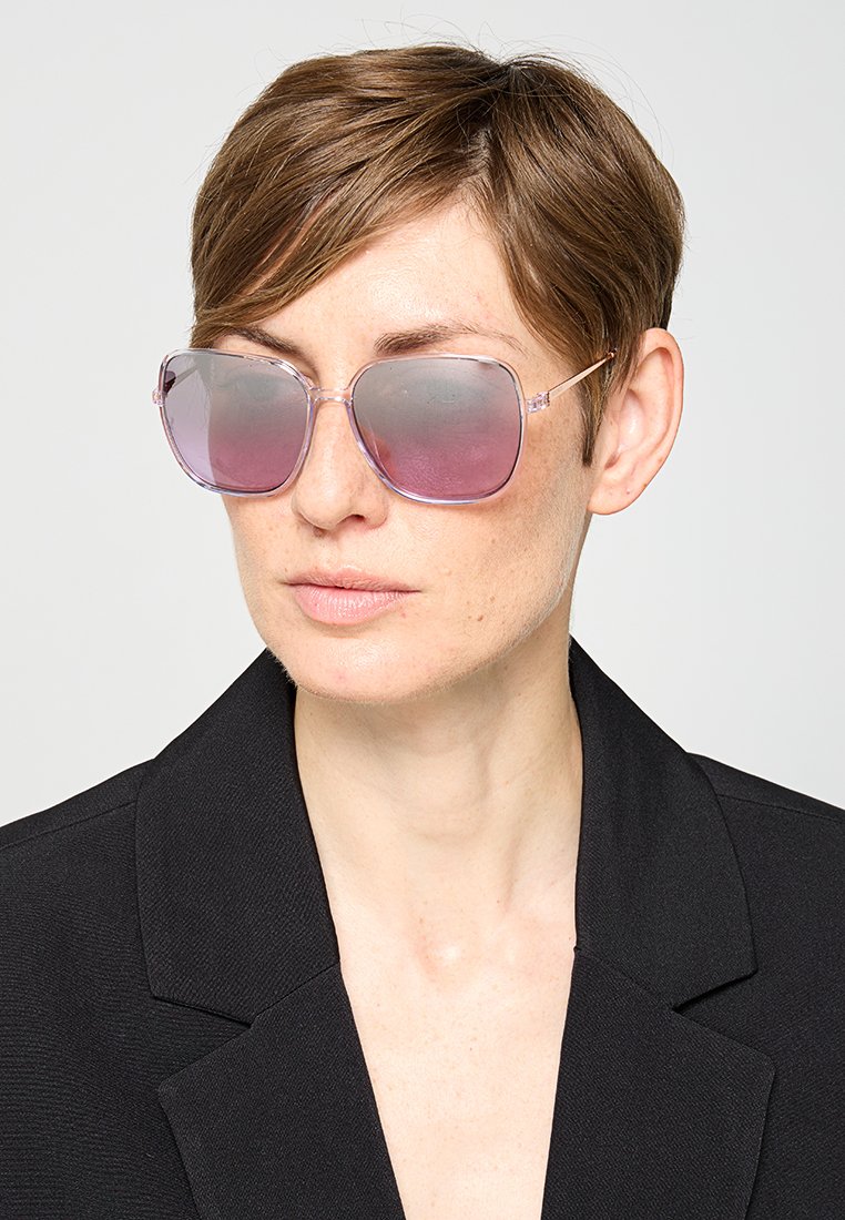 Lila getönte Sonnenbrille mit einem klaren Rahmen, übergroßer geometrischer Form und dünnen Metallbügeln. Getragen von einem Model mit kurzen Haaren.