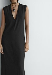 Mustattu hihaton mekko, jossa on syvä V-kaula-aukko, valmistettu sileästä kankaasta. Yhdistetty ohueen mustaan choker-kaulaketjuun, jossa on geometrinen riipus.