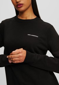 Juodas megztinis iš minkštos medžiagos su apvalia iškirpte ir ilgomis rankovėmis. Krūtinėje yra baltas "KARL LAGERFELD" logotipas.