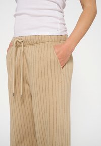 Beige ribcordbroeken met een drawstring taille, voorzien van zijzakken en een subtiele geborduurde logo. Het materiaal lijkt zacht en gestructureerd.