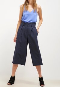 Modrý saténový top na ramínka spojený s tmavě modrými vysokým pasem, kalhotami culottes. Culottes mají sklady a široký zkrácený střih nohavic. Černé boty na podpatku s výřezy.