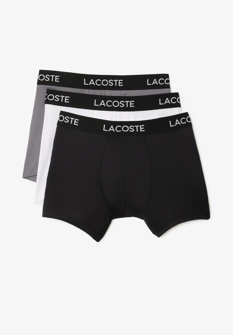 Trois paires de boxers en noir, blanc et gris. Chacune a une ceinture noire avec le logo "LACOSTE" et une texture de tissu lisse.