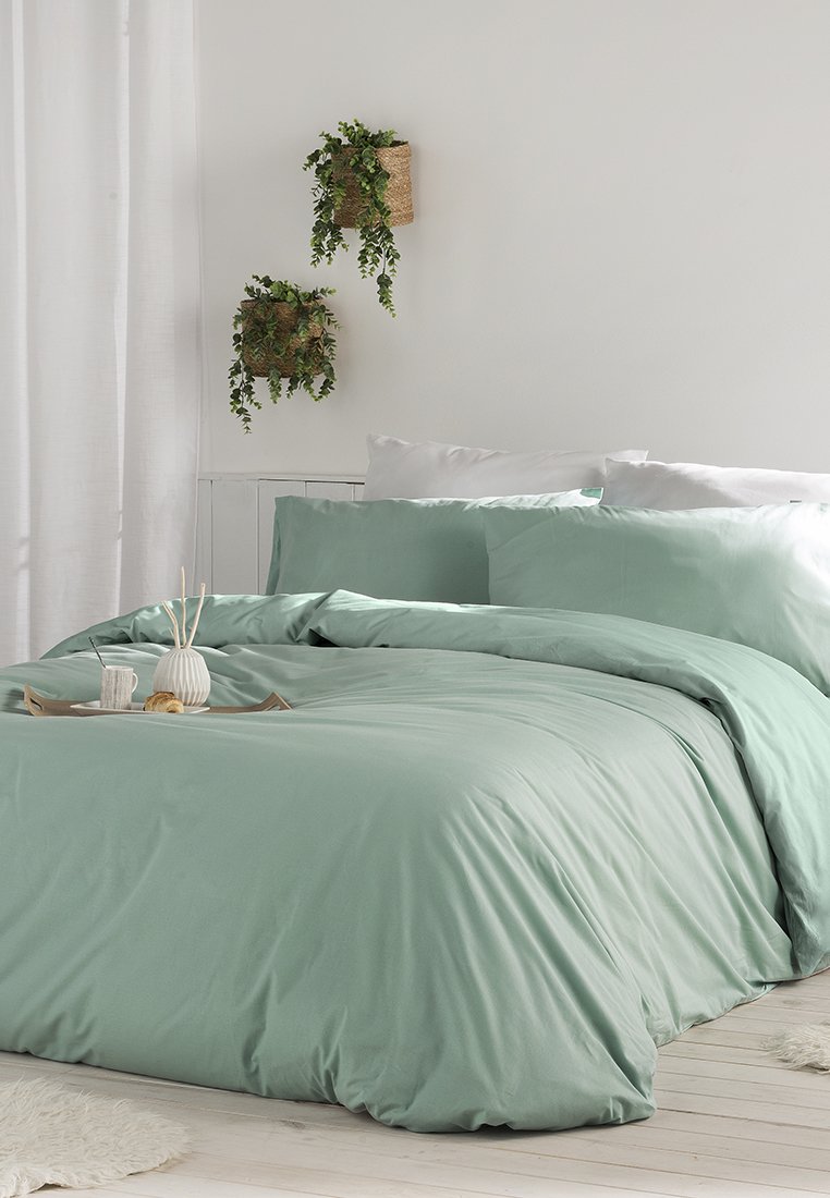 Muntgroen beddengoedset met een gladde, zachte textuur; inclusief kussens en een deken die over een houten bed zijn gedrapeerd. Decoratieve wandplanten erboven.