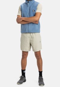 Světle modrá fleecová vesta se zipem, krémová košile s krátkým rukávem, světle béžové šortky, černé ponožky a šedé sportovní boty.