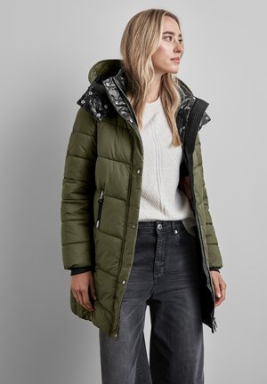 Olijfgroene pufferjas met een ritssluiting aan de voorkant, zwart gemêleerde voering en een afneembare capuchon; gedragen met een crèmekleurige gebreide trui en donkere jeans.