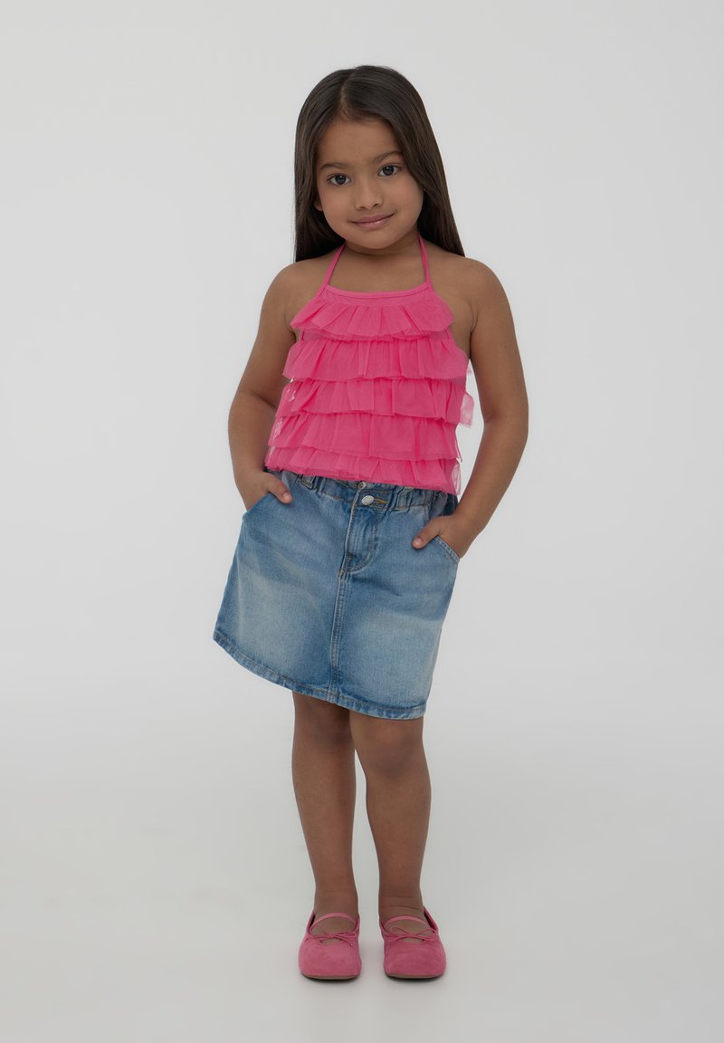 Top halter rosa arricciato con gonna di denim, entrambi stili casual. L'outfit è completato da scarpe piatte rosa, in piedi contro uno sfondo bianco.