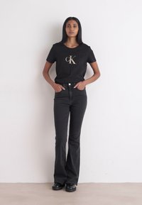 Camiseta de algodão preta com um logótipo dourado, combinada com jeans flare pretos de cintura alta. Modelo de pé, usando sapatos pretos chunky, contra um fundo branco.