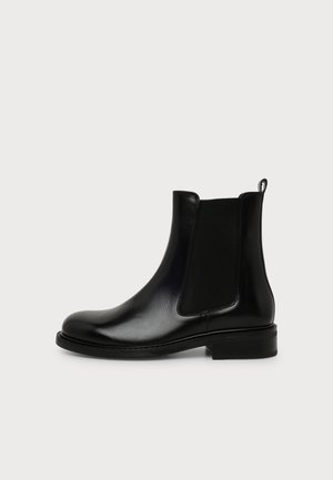 Bottine Chelsea en cuir noir avec un panneau latéral élastique et un talon bas sur un fond clair uni.