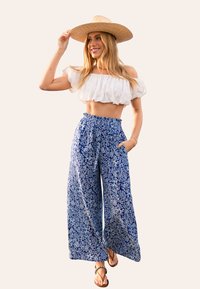 Witte off-the-shoulder crop top gecombineerd met wijde blauwe broek met een bloemenpatroon. Model draagt een strohoed en sandalen.
