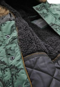 Veste verte avec motif de dinosaure, doublure noire duveteuse, texture matelassée, et accents marron contrastants autour des bords.