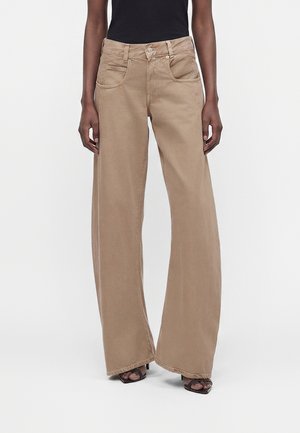 Person iført beige bukser med høj talje og vide ben samt sorte sandaler med hæl, stående mod en ensfarvet lys baggrund.